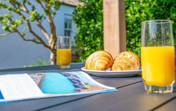 Two glasses of orange juice with croissants and a brochure on a table outdoors.