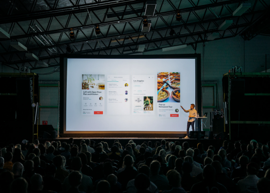 Presenter on stage beside a large screen displaying a digital food menu. Audience viewing.